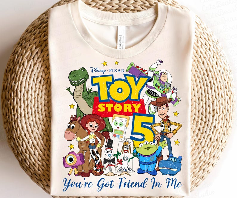 May include: A cream-colored t-shirt featuring a colorful graphic of Disney Pixar Toy Story characters including Woody, Buzz Lightyear, Jessie, Rex, Bullseye, and others. The design includes the text Toy Story 5 and You've Got Friend In Me.