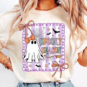 May include: Cream-coloured t-shirt with a Halloween graphic. The design features a white ghost with a bow, a spider web, bats, stars, and the words "Spooky Season" in colourful letters. The graphic is framed with a purple border.