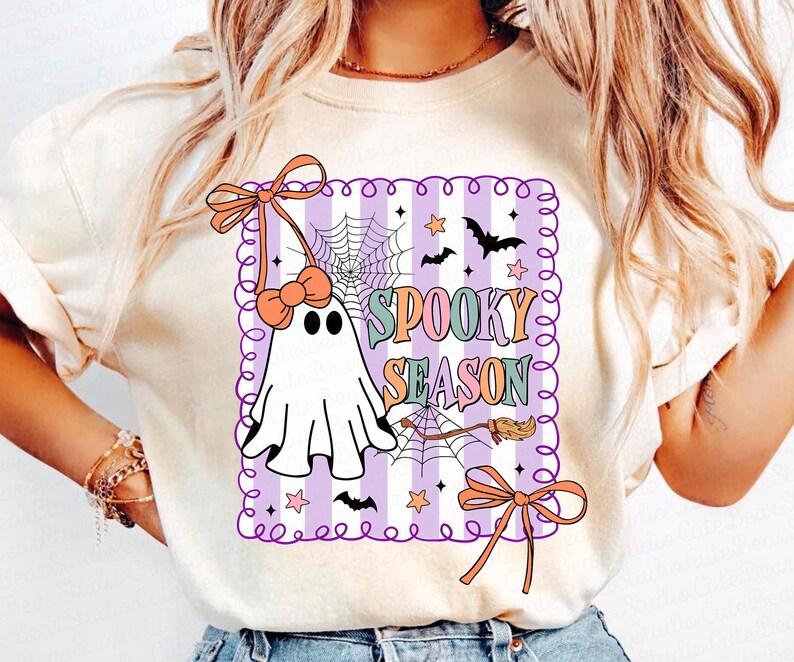 May include: Cream-coloured t-shirt with a Halloween graphic. The design features a white ghost with a bow, a spider web, bats, stars, and the words "Spooky Season" in colourful letters. The graphic is framed with a purple border.