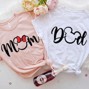May include: Two t-shirts, one blush pink and one white, are displayed. The pink shirt has the word "Mom" in black script with a Mickey Mouse head silhouette and a red bow. The white shirt says "Dad" in black script with a Mickey Mouse head silhouette.