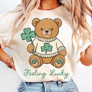 May include: Cream-colored t-shirt featuring a cartoon teddy bear holding a green shamrock. The bear wears a sweater with a shamrock design. The words "Feeling Lucky" are written in green script below the bear. A St. Patrick's Day themed graphic tee.