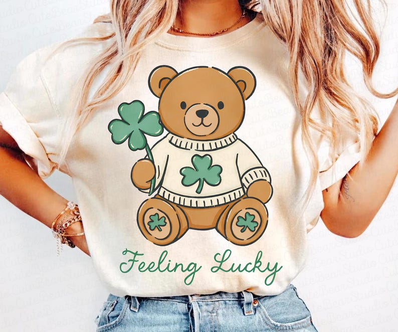 May include: Cream-colored t-shirt featuring a cartoon teddy bear holding a green shamrock. The bear wears a sweater with a shamrock design. The words "Feeling Lucky" are written in green script below the bear. A St. Patrick's Day themed graphic tee.