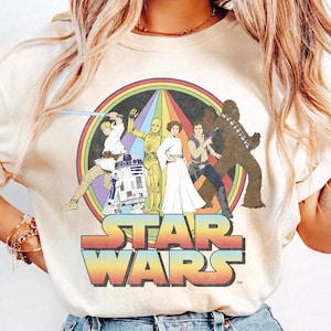 K&ouml;nnte beinhalten: Ein cremefarbenes T-Shirt mit einem Star Wars-Motiv im Vintage-Stil. Das Motiv zeigt Luke Skywalker, Prinzessin Leia, Chewbacca, R2-D2 und C-3PO, die vor einem Regenbogen stehen. Der Text "STAR WARS" ist unter dem Motiv gedruckt.