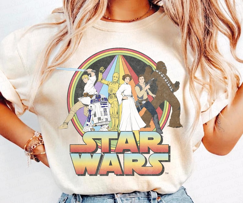 K&ouml;nnte beinhalten: Ein cremefarbenes T-Shirt mit einem Star Wars-Motiv im Vintage-Stil. Das Motiv zeigt Luke Skywalker, Prinzessin Leia, Chewbacca, R2-D2 und C-3PO, die vor einem Regenbogen stehen. Der Text "STAR WARS" ist unter dem Motiv gedruckt.