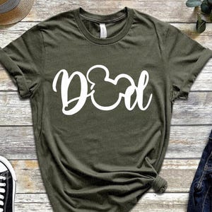 May include: Olive green t-shirt with the word "Dad" in white script, featuring a Mickey Mouse head in place of the "O". The shirt is made of soft material and has rolled-up sleeves. A casual, everyday wear tee.