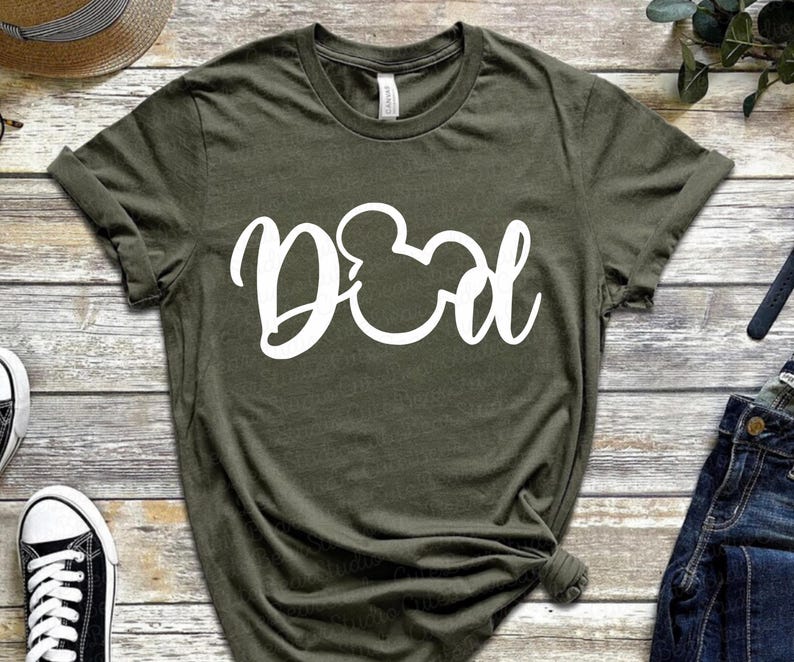 May include: Olive green t-shirt with the word "Dad" in white script, featuring a Mickey Mouse head in place of the "O". The shirt is made of soft material and has rolled-up sleeves. A casual, everyday wear tee.