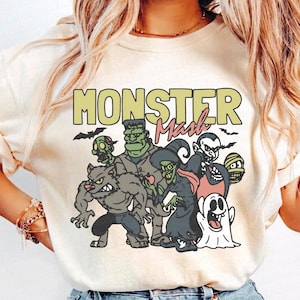 May include: A cream-coloured t-shirt with a graphic of cartoon monsters, such as a werewolf, Frankenstein's monster, a witch, and a vampire. The text "MONSTER Mash" is printed across the top in green and pink, with bats flying above.