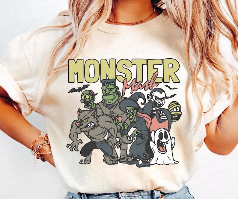 May include: A cream-coloured t-shirt with a graphic of cartoon monsters, such as a werewolf, Frankenstein's monster, a witch, and a vampire. The text "MONSTER Mash" is printed across the top in green and pink, with bats flying above.