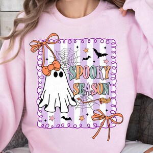 May include: A pale pink sweatshirt with a Halloween design. The graphic features a white ghost, spiderwebs, bats, stars, and the words "Spooky Season" in various colours. The design is framed with a purple border and orange bows.