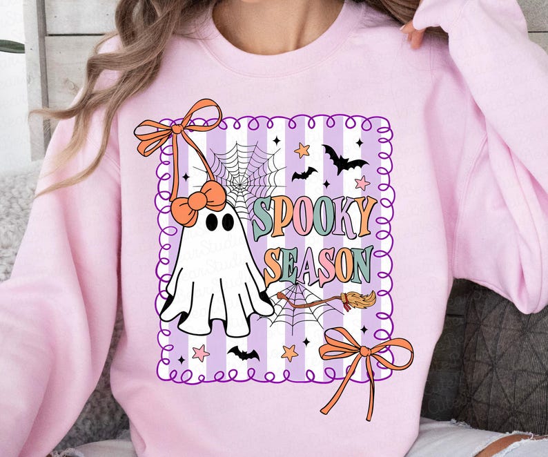 May include: A pale pink sweatshirt with a Halloween design. The graphic features a white ghost, spiderwebs, bats, stars, and the words "Spooky Season" in various colours. The design is framed with a purple border and orange bows.
