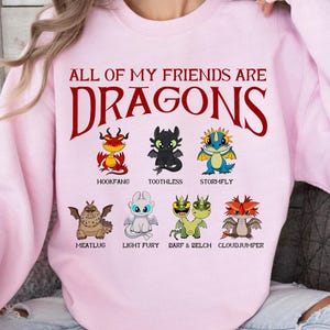 K&ouml;nnte beinhalten: Hellrosa Sweatshirt mit dem roten Schriftzug "ALL OF MY FRIENDS ARE DRAGONS". Darunter befinden sich Cartoon-Drachen, darunter Hookfang und Toothless. Das Sweatshirt hat einen Rundhalsausschnitt.
