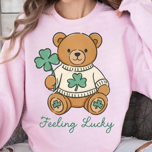 May include: A light pink sweatshirt featuring a cartoon teddy bear holding a green shamrock. The bear wears a white sweater with a shamrock design. The words "Feeling Lucky" are written in green script below the bear.