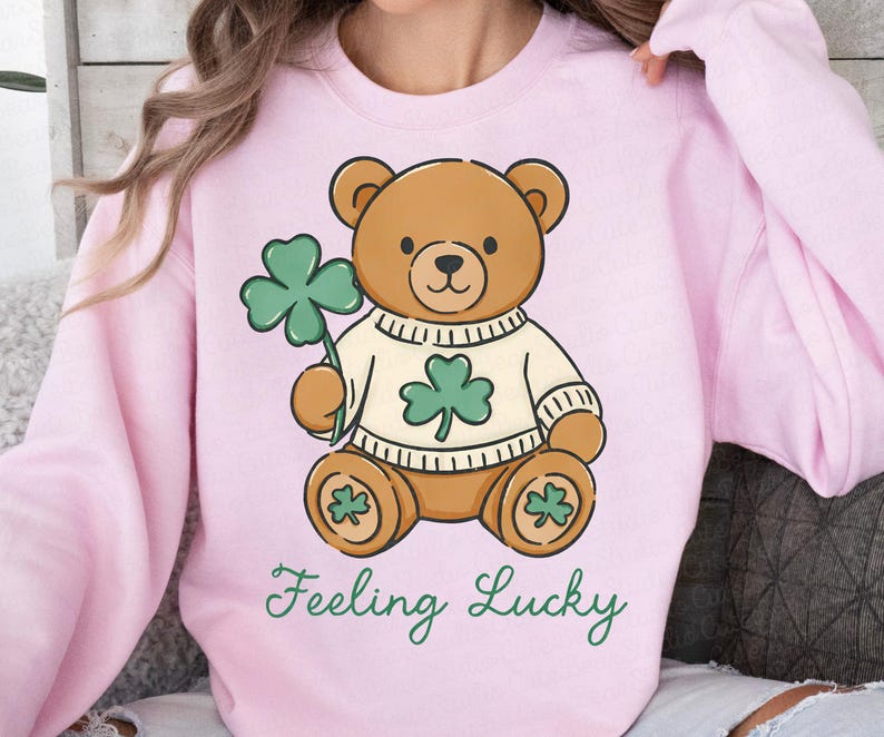 May include: A light pink sweatshirt featuring a cartoon teddy bear holding a green shamrock. The bear wears a white sweater with a shamrock design. The words "Feeling Lucky" are written in green script below the bear.