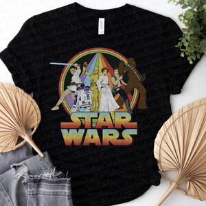 K&ouml;nnte beinhalten: Schwarzes T-Shirt mit einer Retro-Star-Wars-Grafik mit Charakteren aus der Original-Trilogie. Die Grafik zeigt Luke Skywalker, Prinzessin Leia, Han Solo, Chewbacca, R2-D2 und C-3PO. Der Text "STAR WARS" ist in einer Regenbogen-Schriftart.
