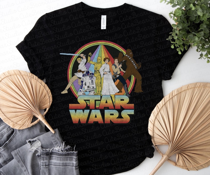 K&ouml;nnte beinhalten: Schwarzes T-Shirt mit einer Retro-Star-Wars-Grafik mit Charakteren aus der Original-Trilogie. Die Grafik zeigt Luke Skywalker, Prinzessin Leia, Han Solo, Chewbacca, R2-D2 und C-3PO. Der Text "STAR WARS" ist in einer Regenbogen-Schriftart.