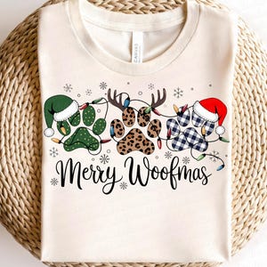 May include: Cream-colored t-shirt with a festive design featuring paw prints decorated with Christmas hats, antlers, and string lights. The text "Merry Woofmas" is written in a script font, surrounded by snowflakes.
