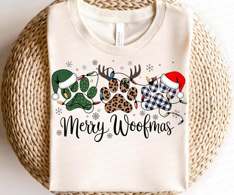 May include: Cream-colored t-shirt with a festive design featuring paw prints decorated with Christmas hats, antlers, and string lights. The text "Merry Woofmas" is written in a script font, surrounded by snowflakes.