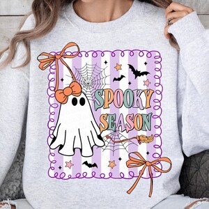 May include: A light grey sweatshirt with a Halloween-themed design. The design features a ghost, bats, stars, a spiderweb, and the words "Spooky Season" in pastel colours. The design is framed with a purple border and orange bows.