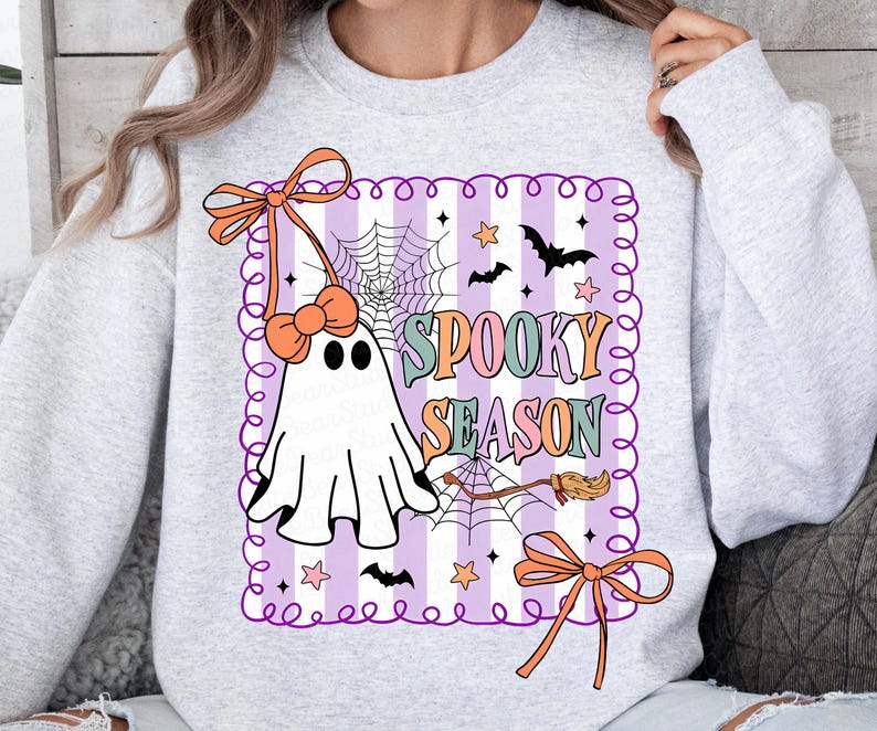 May include: A light grey sweatshirt with a Halloween-themed design. The design features a ghost, bats, stars, a spiderweb, and the words "Spooky Season" in pastel colours. The design is framed with a purple border and orange bows.