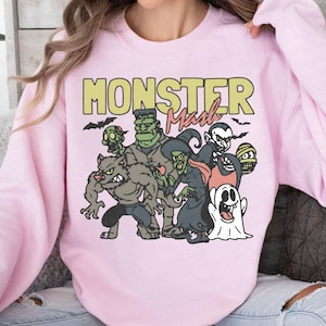 May include: A pale pink sweatshirt with a cartoon illustration of classic monsters such as a werewolf, Frankenstein's monster, a witch, and a vampire. The words "MONSTER Mash" are printed above the characters in yellow and black.