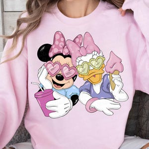 May include: Pink sweatshirt featuring Minnie Mouse and Daisy Duck wearing heart-shaped sunglasses. Minnie holds a drink, and Daisy holds an ice cream cone. Both characters are wearing bows. The sweatshirt is a light pink color.