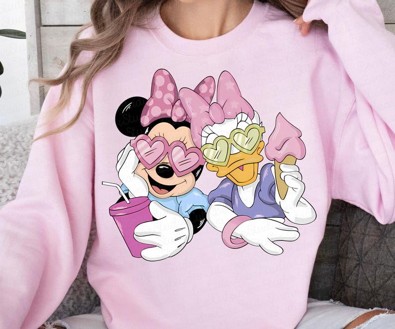 May include: Pink sweatshirt featuring Minnie Mouse and Daisy Duck wearing heart-shaped sunglasses. Minnie holds a drink, and Daisy holds an ice cream cone. Both characters are wearing bows. The sweatshirt is a light pink color.