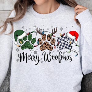 May include: A light gray sweatshirt with a festive design. The design features paw prints decorated with a green elf hat, reindeer antlers, and a Santa hat. The words "Merry Woofmas" are written in a script font, with snowflakes scattered around the design.