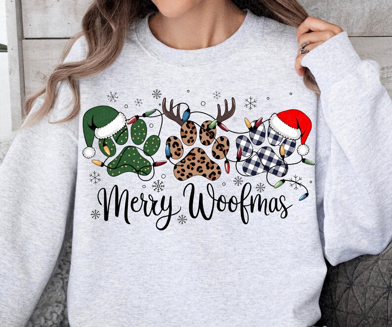 May include: A light gray sweatshirt with a festive design. The design features paw prints decorated with a green elf hat, reindeer antlers, and a Santa hat. The words "Merry Woofmas" are written in a script font, with snowflakes scattered around the design.