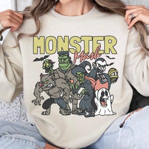 May include: Beige sweatshirt featuring a cartoon illustration of classic monsters such as a werewolf, Frankenstein's monster, and a vampire. The words "MONSTER Mash" are printed above the characters in yellow and red.