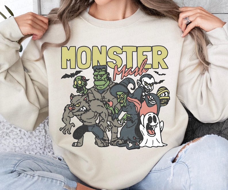May include: Beige sweatshirt featuring a cartoon illustration of classic monsters such as a werewolf, Frankenstein's monster, and a vampire. The words "MONSTER Mash" are printed above the characters in yellow and red.