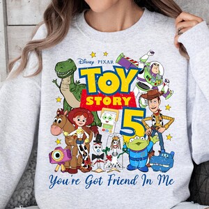 May include: A light gray crewneck sweatshirt featuring a colorful Disney Pixar Toy Story 5 graphic. The design includes characters like Woody, Buzz Lightyear, Jessie, Rex, and the Alien, with the text Toy Story 5 and You've Got Friend In Me.