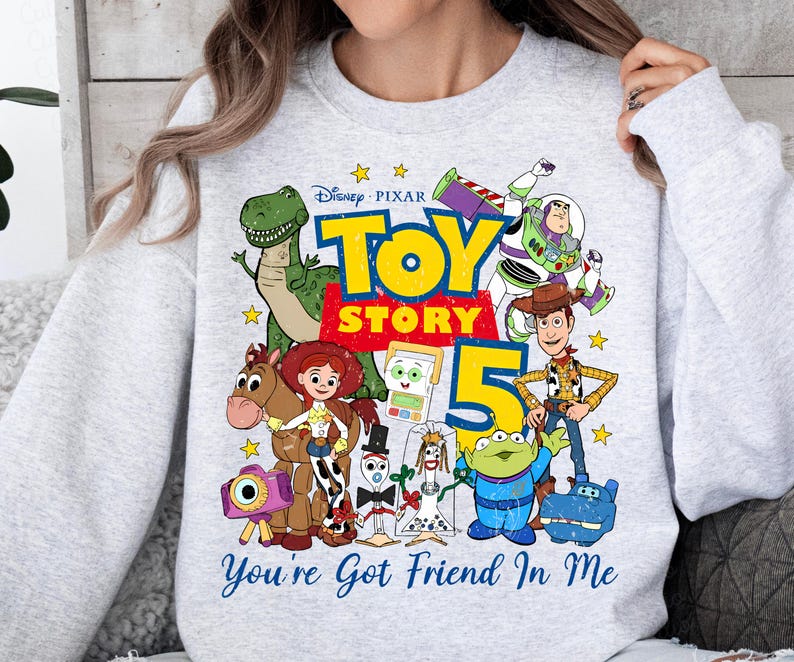May include: A light gray crewneck sweatshirt featuring a colorful Disney Pixar Toy Story 5 graphic. The design includes characters like Woody, Buzz Lightyear, Jessie, Rex, and the Alien, with the text Toy Story 5 and You've Got Friend In Me.