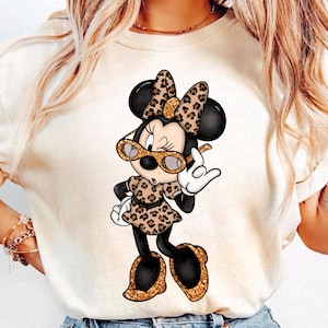 Puede incluir: Una camiseta blanca con una imagen de dibujos animados de Minnie Mouse con gafas de sol de estampado de leopardo y un vestido de estampado de leopardo. Ella está levantando la mano en señal de paz.