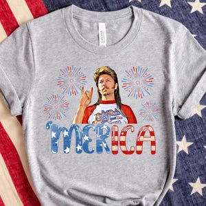 May include: Grey t-shirt featuring a graphic of a man with long blonde hair and a beard wearing a red, white and blue shirt with the words "Rock N Roll" on it. The graphic is surrounded by red, white and blue fireworks. The text "'MERICA" is below the graphic in red, white and blue.