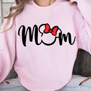 May include: A light pink sweatshirt with the word "Mom" in black script. The "O" in "Mom" is replaced with a Mickey Mouse head silhouette, and a red bow is on top.