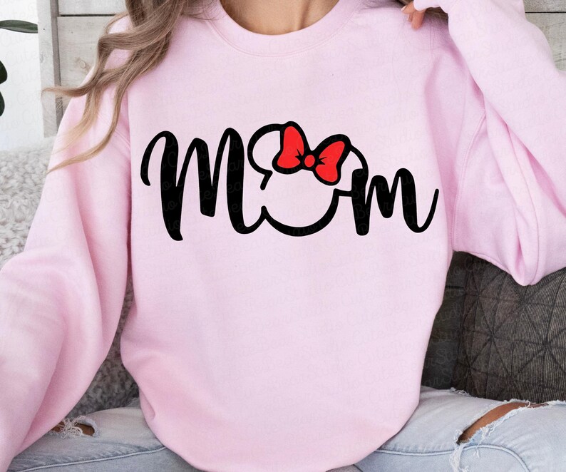 May include: A light pink sweatshirt with the word "Mom" in black script. The "O" in "Mom" is replaced with a Mickey Mouse head silhouette, and a red bow is on top.