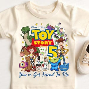 May include: A cream-colored short-sleeve t-shirt featuring a colorful graphic of Toy Story characters including Woody, Buzz Lightyear, Jessie, Rex, and others. The design includes the text "Disney Pixar TOY STORY 5" and "You're Got Friend In Me".