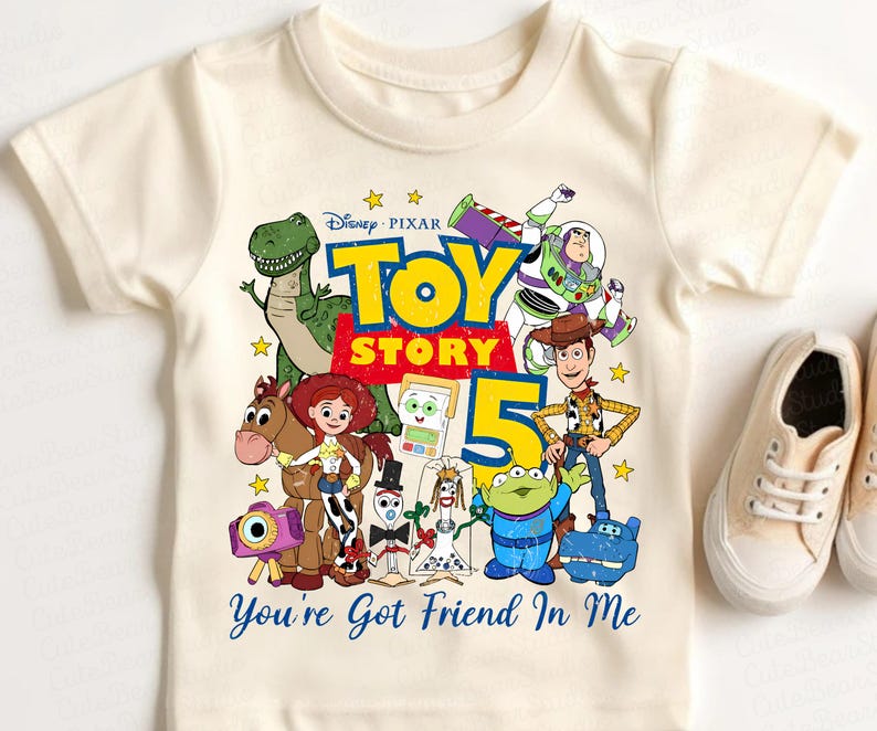 May include: A cream-colored short-sleeve t-shirt featuring a colorful graphic of Toy Story characters including Woody, Buzz Lightyear, Jessie, Rex, and others. The design includes the text "Disney Pixar TOY STORY 5" and "You're Got Friend In Me".