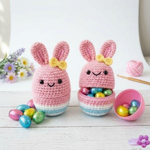 Peut inclure: Deux porte-œufs en forme de lapin rose au crochet avec des nœuds jaunes, des yeux noirs et des visages souriants. L'un est ouvert, révélant des œufs en chocolat emballés dans du papier d'aluminium. Décoration de Pâques.