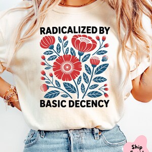 Pode incluir: T-shirt creme com um design floral vermelho e azul e o texto "RADICALIZED BY BASIC DECENCY". O design inclui flores e folhas estilizadas. A camisa é feita de um material macio.