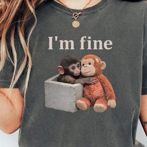 Puede incluir: Camiseta gris oscuro jaspeado con el texto "I'm fine" sobre una ilustración de dos monos abrazándose. Un mono está en una caja gris y el otro lo abraza. La camiseta es de color gris oscuro.