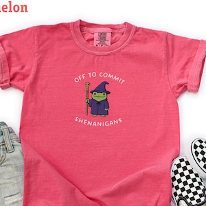 Può includere: Una maglietta da ragazzo color anguria con maniche arrotolate, con un cartone animato di una rana mago con la scritta "OFF TO COMMIT SHENANIGANS". La maglietta è abbinata a pantaloncini di jeans e scarpe slip-on a scacchi bianchi e neri.