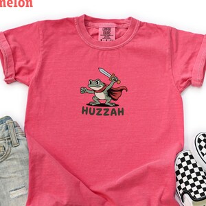 Può includere: Maglietta da bambino color anguria con un cartone animato di una rana che tiene una spada e indossa un mantello. La parola "Huzzah" è sotto. L'immagine include pantaloncini di jeans e scarpe slip-on a scacchi bianchi e neri.
