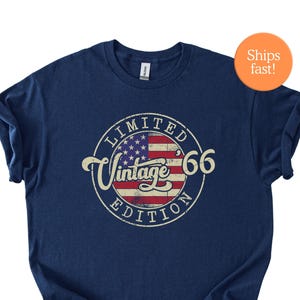 Può includere: T-shirt blu navy con grafica invecchiata raffigurante la bandiera americana e le parole "Vintage 66 Limited Edition" in un design circolare. La maglietta ha un girocollo classico e maniche arrotolate.