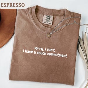 Può includere: T-shirt color espresso con la scritta bianca "sorry, I can't. I have a couch commitment". La maglietta ha girocollo e maniche corte. Una collana d'argento con pendente è drappeggiata sulla maglietta.