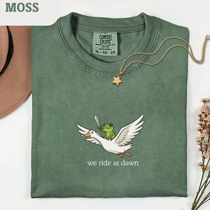 Può includere: Una maglietta verde muschio con una grafica stravagante di una rana che cavalca un'oca, che tiene una spada, con la scritta "we ride at dawn". Una collana con pendente a stella dorata è appoggiata sulla maglietta. La maglietta è etichettata XL-TG-EG.