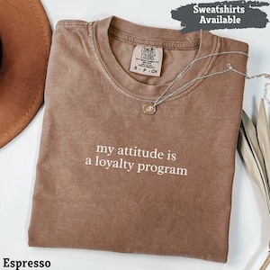 Può includere: T-shirt color espresso con la scritta bianca "my attitude is a loyalty program". La maglietta ha girocollo e maniche corte. Una collana d'argento con un ciondolo è drappeggiata sulla maglietta. La maglietta è piegata.