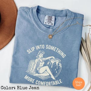 Può includere: T-shirt Comfort Colors blu jeans con la scritta "Slip Into Something More Comfortable" e un'illustrazione in stile vintage. Una collana d'oro con un ciondolo è appoggiata sulla maglietta. La maglietta è una taglia piccola.