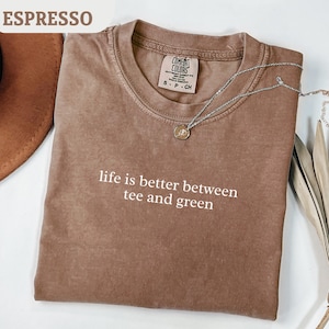 Può includere: T-shirt color espresso con la scritta bianca "life is better between tee and green". Una collana d'argento con un ciondolo è appoggiata sulla maglietta. La maglietta è del marchio Comfort Colors.