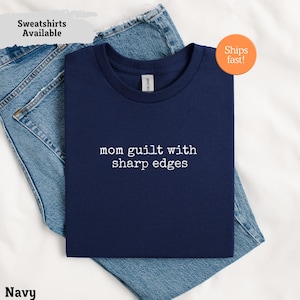 Può includere: T-shirt blu navy con la scritta bianca "mom guilt with sharp edges". La maglietta è piegata sopra dei jeans blu. Le parole "Sweatshirts Available" sono in un riquadro grigio chiaro. Un adesivo arancione dice "Ships fast!"